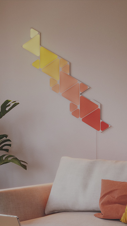 此圖片顯示客廳沙發上方牆壁安裝的 Nanoleaf Shapes 三角形和迷你三角形智能燈板佈局。 模組化的變色智能燈板由連接片一併連接，且有着超過 1600 萬種顏色。
