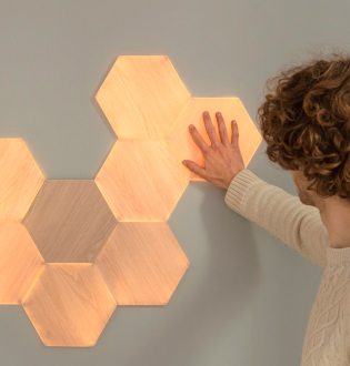 此圖片展示牆壁的 Nanoleaf Elements 木紋六角形智能燈板。 智能燈板對觸摸和音樂有反應，並讓您的家居充滿柔和的照明。 輕觸木質外觀面板或播放您喜愛的音樂，讓您的燈飾散發流動的光線。
