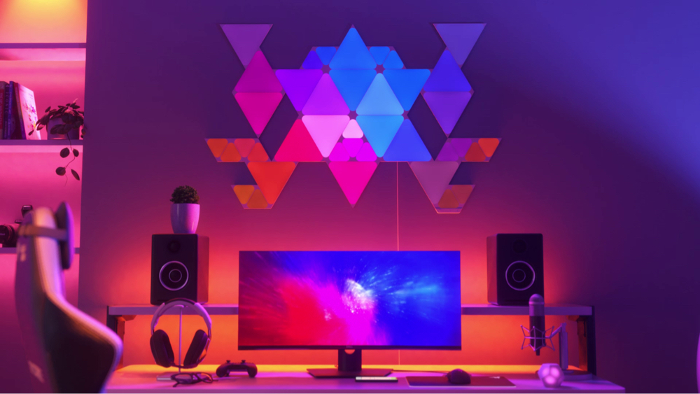 ภาพแผ่นไฟรูปสี่เหลี่ยมรุ่น Nanoleaf Shapes Triangles และ Mini Triangles บนผนังเหนือโต๊ะทำงาน หลังจอแสดงผลในห้องเล่นเกม ไฟ RGB เหล่านี้มีสีให้เลือกมากกว่า 16 ล้านสี เหมาะสำหรับเกมเมอร์ในบ้านของคุณ แผ่นไฟสมาร์ทไลต์แบบโมดูลที่เชื่อมต่อกันด้วยตัวเชื่อมเพื่อสร้างงานดีไซน์